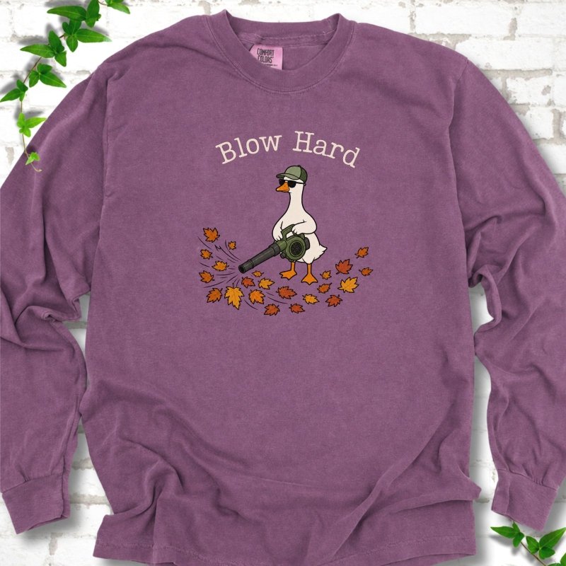 Berry long sleeve T-shirt flat lay with ivory "Blow Hard" lettering above a white goose in a green cap and sunglasses using a leaf blower on a pile of orange and gold fall leaves, on white brick with ivy sprigs.