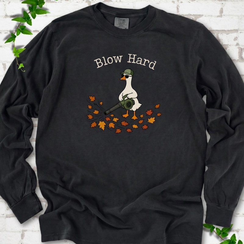 Black long sleeve T-shirt flat lay with ivory "Blow Hard" lettering above a white goose in a green cap and sunglasses using a leaf blower on a pile of orange and gold fall leaves, on white brick with ivy sprigs.