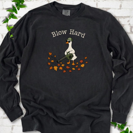 Black long sleeve T-shirt flat lay with ivory "Blow Hard" lettering above a white goose in a green cap and sunglasses using a leaf blower on a pile of orange and gold fall leaves, on white brick with ivy sprigs.
