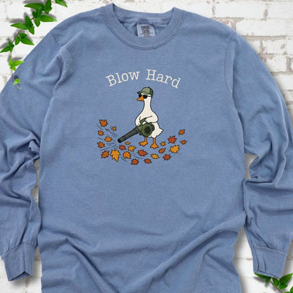 Blue Jean long sleeve T-shirt flat lay with ivory "Blow Hard" lettering above a white goose in a green cap and sunglasses using a leaf blower on a pile of orange and gold fall leaves, on white brick with ivy sprigs.
