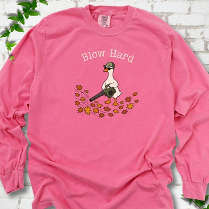 Crunchberry long sleeve T-shirt flat lay with ivory "Blow Hard" lettering above a white goose in a green cap and sunglasses using a leaf blower on a pile of orange and gold fall leaves, on white brick with ivy sprigs.
