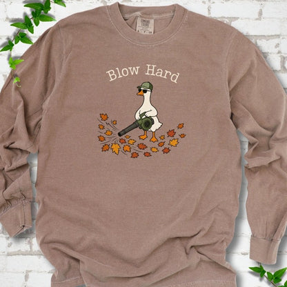 Espresso long sleeve T-shirt flat lay with ivory "Blow Hard" lettering above a white goose in a green cap and sunglasses using a leaf blower on a pile of orange and gold fall leaves, on white brick with ivy sprigs.