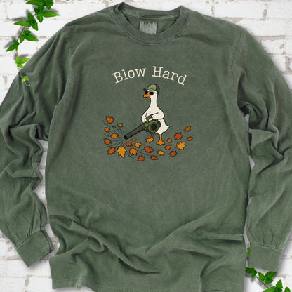Hemp long sleeve T-shirt flat lay with ivory "Blow Hard" lettering above a white goose in a green cap and sunglasses using a leaf blower on a pile of orange and gold fall leaves, on white brick with ivy sprigs.