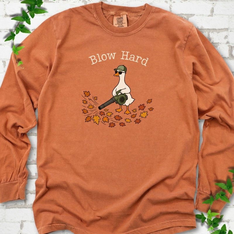Yam long sleeve T-shirt flat lay with ivory "Blow Hard" lettering above a white goose in a green cap and sunglasses using a leaf blower on a pile of orange and gold fall leaves, on white brick with ivy sprigs.