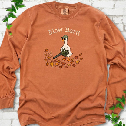 Yam long sleeve T-shirt flat lay with ivory "Blow Hard" lettering above a white goose in a green cap and sunglasses using a leaf blower on a pile of orange and gold fall leaves, on white brick with ivy sprigs.