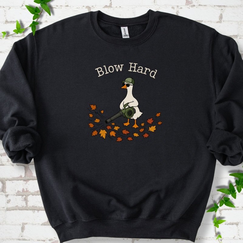 Black sweatshirt flat lay with ivory "Blow Hard" lettering above a white goose in a green cap and sunglasses using a leaf blower on a pile of orange and gold fall leaves, on white brick with ivy sprigs.