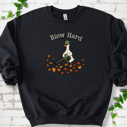 Black sweatshirt flat lay with ivory "Blow Hard" lettering above a white goose in a green cap and sunglasses using a leaf blower on a pile of orange and gold fall leaves, on white brick with ivy sprigs.