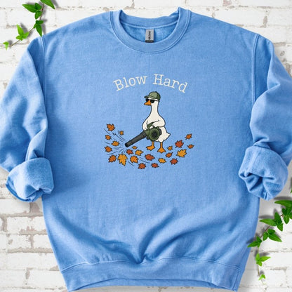 Carolina Blue sweatshirt flat lay with ivory "Blow Hard" lettering above a white goose in a green cap and sunglasses using a leaf blower on a pile of orange and gold fall leaves, on white brick with ivy sprigs.