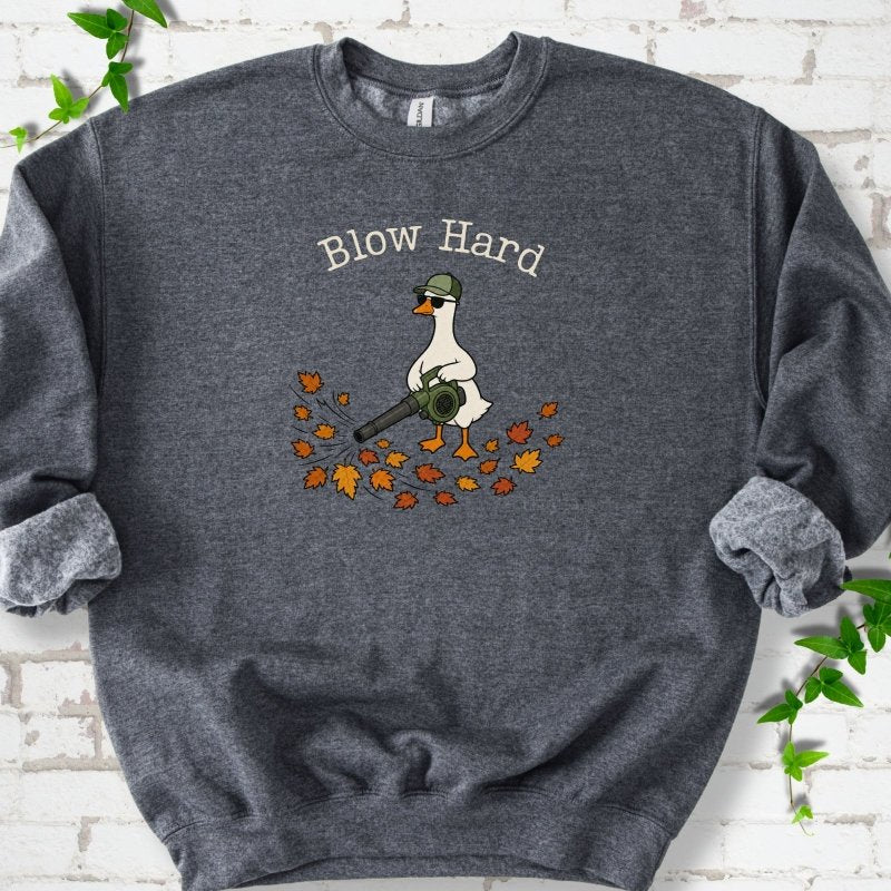 Dark Heather sweatshirt flat lay with ivory "Blow Hard" lettering above a white goose in a green cap and sunglasses using a leaf blower on a pile of orange and gold fall leaves, on white brick with ivy sprigs.