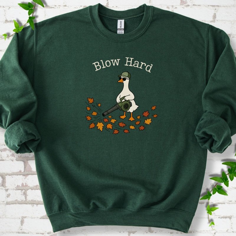 Forest Green sweatshirt flat lay with ivory "Blow Hard" lettering above a white goose in a green cap and sunglasses using a leaf blower on a pile of orange and gold fall leaves, on white brick with ivy sprigs.
