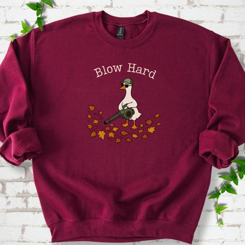 Maroon sweatshirt flat lay with ivory "Blow Hard" lettering above a white goose in a green cap and sunglasses using a leaf blower on a pile of orange and gold fall leaves, on white brick with ivy sprigs.