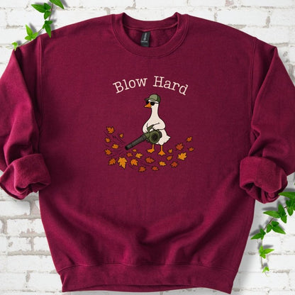 Maroon sweatshirt flat lay with ivory "Blow Hard" lettering above a white goose in a green cap and sunglasses using a leaf blower on a pile of orange and gold fall leaves, on white brick with ivy sprigs.