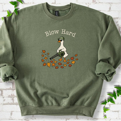 Military Green sweatshirt flat lay with ivory "Blow Hard" lettering above a white goose in a green cap and sunglasses using a leaf blower on a pile of orange and gold fall leaves, on white brick with ivy sprigs.
