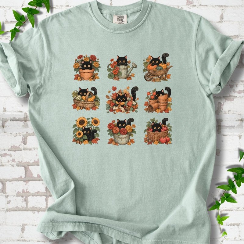 Front flat lay of a Bay short sleeve T-shirt with a nine-panel grid of black cats among fall garden flowers, pots and leaves, on white brick with green leafy sprigs.