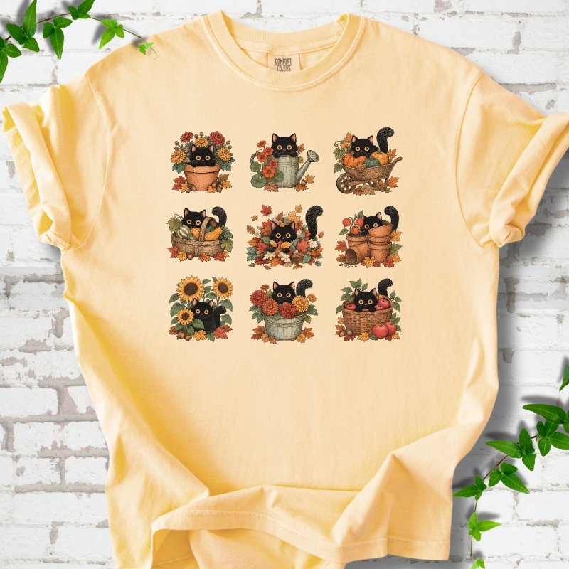 Front flat lay of a Butter short sleeve T-shirt with a nine-panel grid of black cats among fall garden flowers, pots and leaves, on white brick with green leafy sprigs.