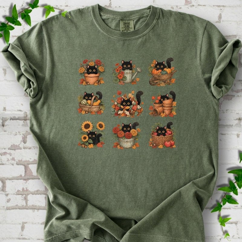 Front flat lay of a Moss short sleeve T-shirt with a nine-panel grid of black cats among fall garden flowers, pots and leaves, on white brick with green leafy sprigs.