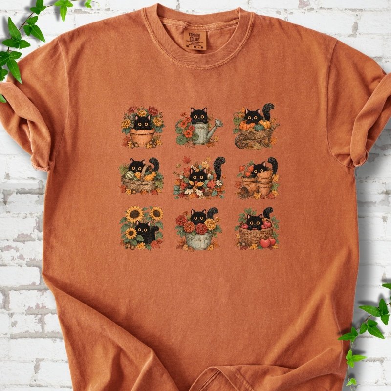 Front flat lay of a Yam short sleeve T-shirt with a nine-panel grid of black cats among fall garden flowers, pots and leaves, on white brick with green leafy sprigs.