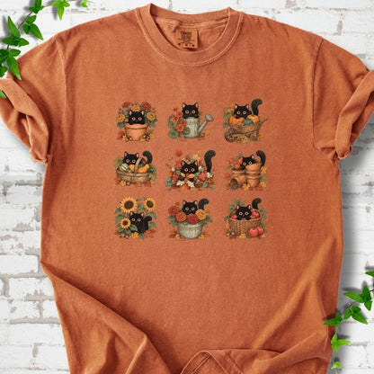 Front flat lay of a Yam short sleeve T-shirt with a nine-panel grid of black cats among fall garden flowers, pots and leaves, on white brick with green leafy sprigs.