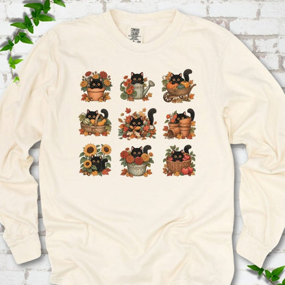Front flat lay of a Ivory long sleeve T-shirt with a nine-panel grid of black cats among fall garden flowers, pots and leaves, on white brick with green leafy sprigs.