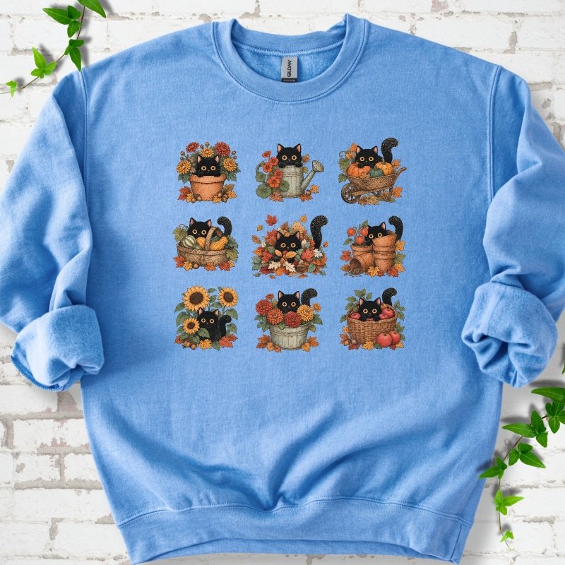 Front flat lay of a Carolina Blue crewneck sweatshirt with a nine-panel grid of black cats among fall garden flowers, pots and leaves, on white brick with green leafy sprigs.