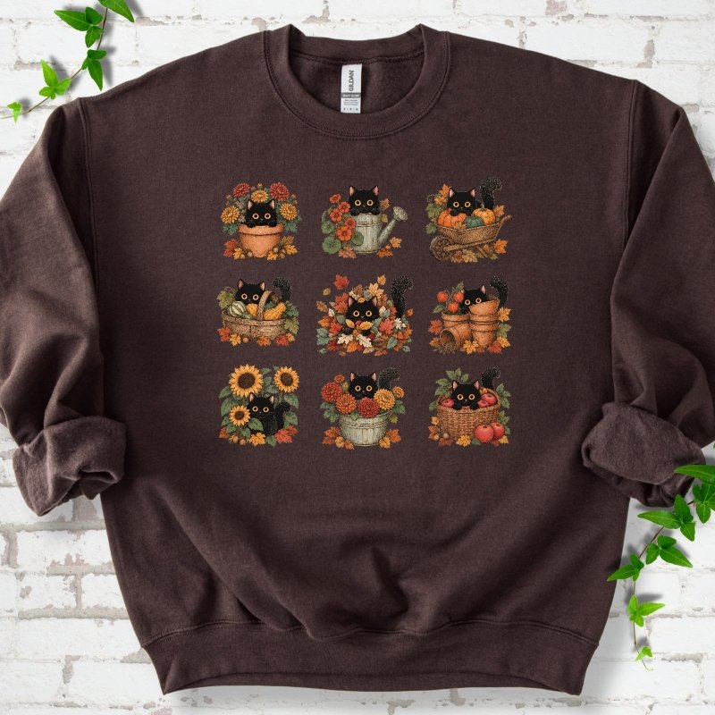 Front flat lay of a Dark Chocolate crewneck sweatshirt with a nine-panel grid of black cats among fall garden flowers, pots and leaves, on white brick with green leafy sprigs.