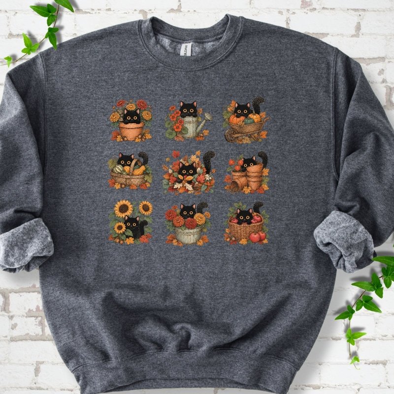 Front flat lay of a Dark Heather crewneck sweatshirt with a nine-panel grid of black cats among fall garden flowers, pots and leaves, on white brick with green leafy sprigs.