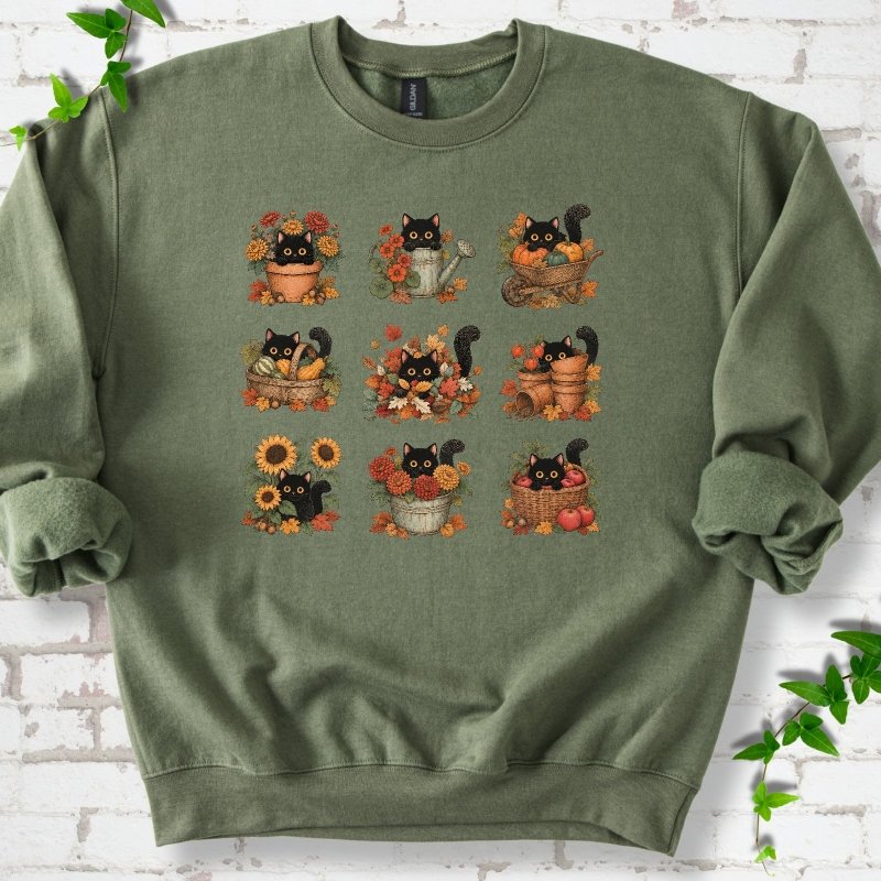 Front flat lay of a Military Green crewneck sweatshirt with a nine-panel grid of black cats among fall garden flowers, pots and leaves, on white brick with green leafy sprigs.