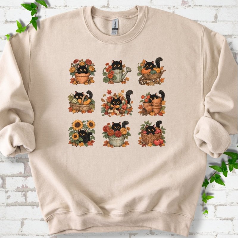 Front flat lay of a Sand crewneck sweatshirt with a nine-panel grid of black cats among fall garden flowers, pots and leaves, on white brick with green leafy sprigs.