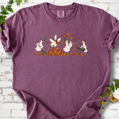 Berry T-shirt flat lay with a row of four white cartoon geese doing fall yard work, one raking in a green cap and sunglasses, one leaping into a pile of orange and gold leaves in a plaid scarf, one in a green beanie tossing leaves and one in an orange beanie using a leaf blower, on white brick with ivy sprigs and an orange butterfly.