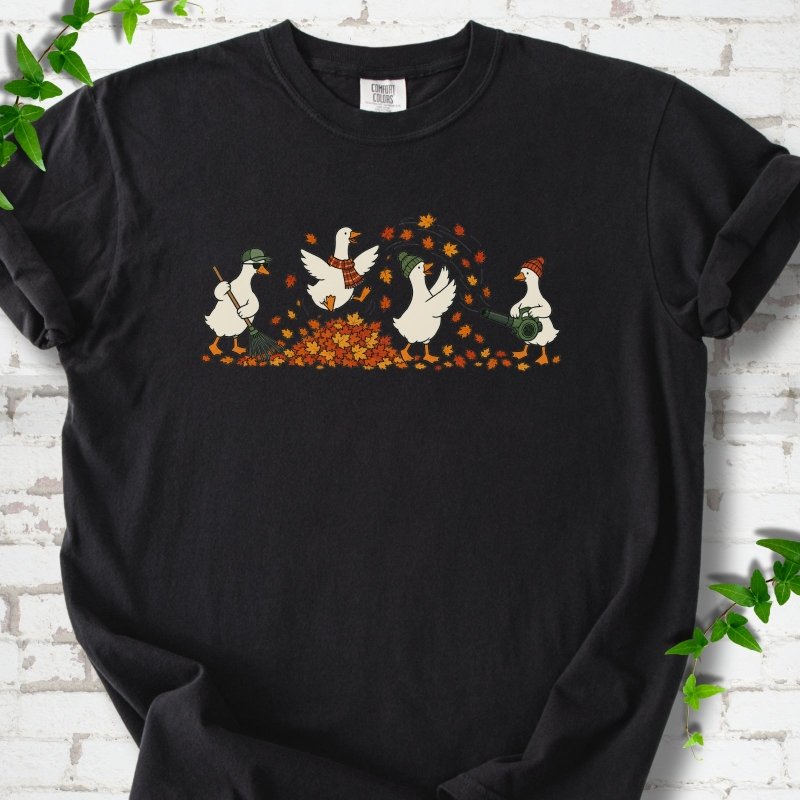 Black T-shirt flat lay with a row of four white cartoon geese doing fall yard work, one raking in a green cap and sunglasses, one leaping into a pile of orange and gold leaves in a plaid scarf, one in a green beanie tossing leaves and one in an orange beanie using a leaf blower, on white brick with ivy sprigs.