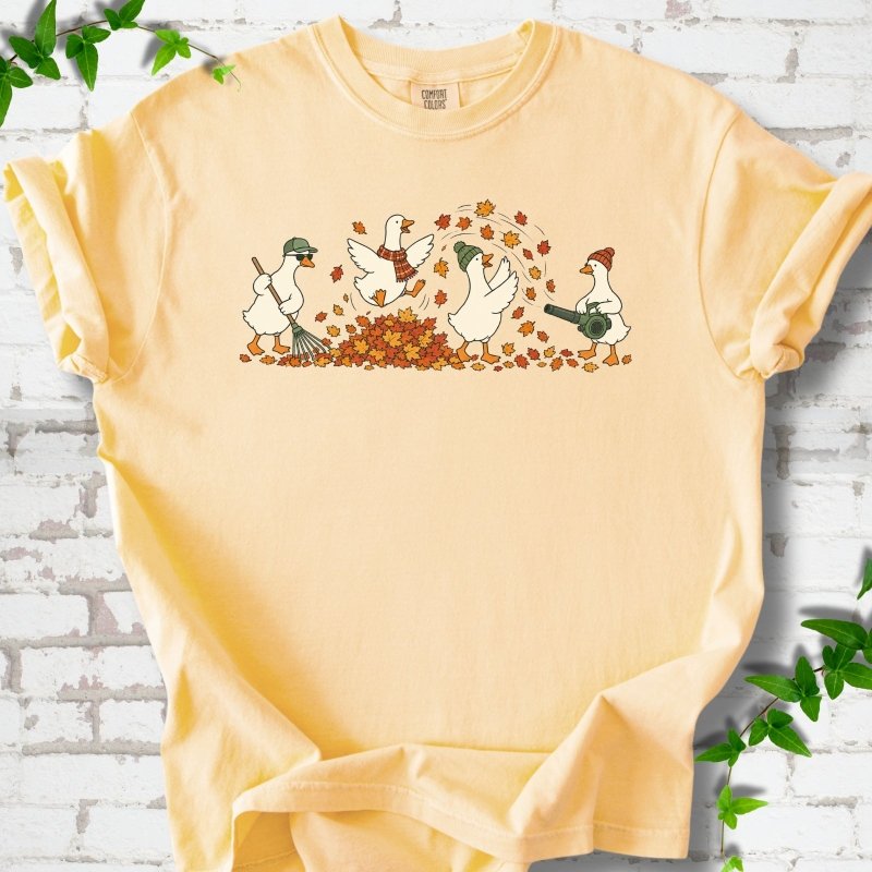 Butter T-shirt flat lay with a row of four white cartoon geese doing fall yard work, one raking in a green cap and sunglasses, one leaping into a pile of orange and gold leaves in a plaid scarf, one in a green beanie tossing leaves and one in an orange beanie using a leaf blower, on white brick with ivy sprigs.