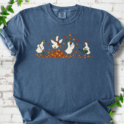Denim T-shirt flat lay with a row of four white cartoon geese doing fall yard work, one raking in a green cap and sunglasses, one leaping into a pile of orange and gold leaves in a plaid scarf, one in a green beanie tossing leaves and one in an orange beanie using a leaf blower, on white brick with ivy sprigs.