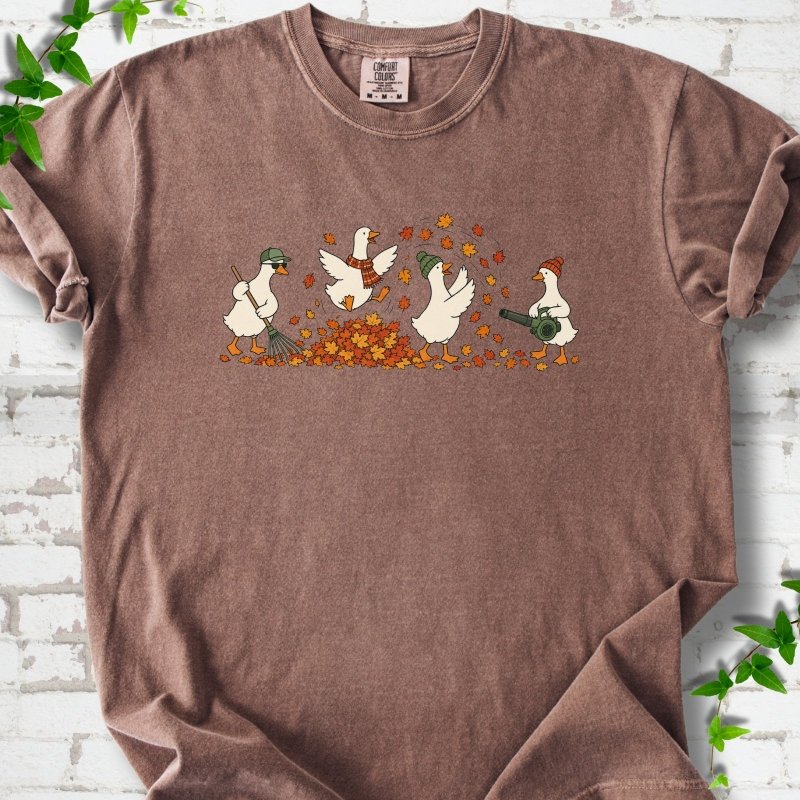Espresso T-shirt flat lay with a row of four white cartoon geese doing fall yard work, one raking in a green cap and sunglasses, one leaping into a pile of orange and gold leaves in a plaid scarf, one in a green beanie tossing leaves and one in an orange beanie using a leaf blower, on white brick with ivy sprigs.