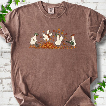 Espresso T-shirt flat lay with a row of four white cartoon geese doing fall yard work, one raking in a green cap and sunglasses, one leaping into a pile of orange and gold leaves in a plaid scarf, one in a green beanie tossing leaves and one in an orange beanie using a leaf blower, on white brick with ivy sprigs.