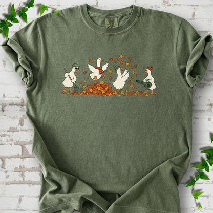 Moss T-shirt flat lay with a row of four white cartoon geese doing fall yard work, one raking in a green cap and sunglasses, one leaping into a pile of orange and gold leaves in a plaid scarf, one in a green beanie tossing leaves and one in an orange beanie using a leaf blower, on white brick with ivy sprigs.