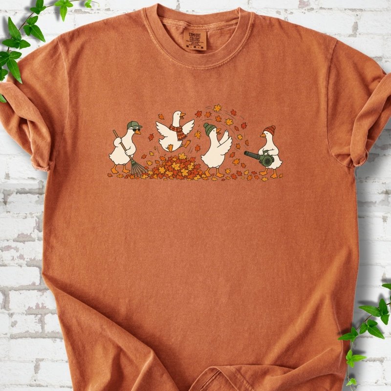 Yam T-shirt flat lay with a row of four white cartoon geese doing fall yard work, one raking in a green cap and sunglasses, one leaping into a pile of orange and gold leaves in a plaid scarf, one in a green beanie tossing leaves and one in an orange beanie using a leaf blower, on white brick with ivy sprigs.