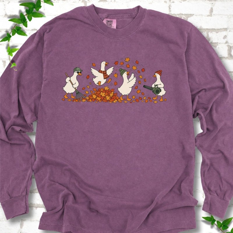 Berry long sleeve T-shirt flat lay with a row of four white cartoon geese doing fall yard work, one raking in a green cap and sunglasses, one leaping into a pile of orange and gold leaves in a plaid scarf, one in a green beanie tossing leaves and one in an orange beanie using a leaf blower, on white brick with ivy sprigs.