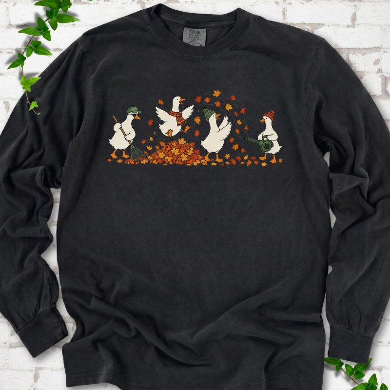 Black long sleeve T-shirt flat lay with a row of four white cartoon geese doing fall yard work, one raking in a green cap and sunglasses, one leaping into a pile of orange and gold leaves in a plaid scarf, one in a green beanie tossing leaves and one in an orange beanie using a leaf blower, on white brick with ivy sprigs.