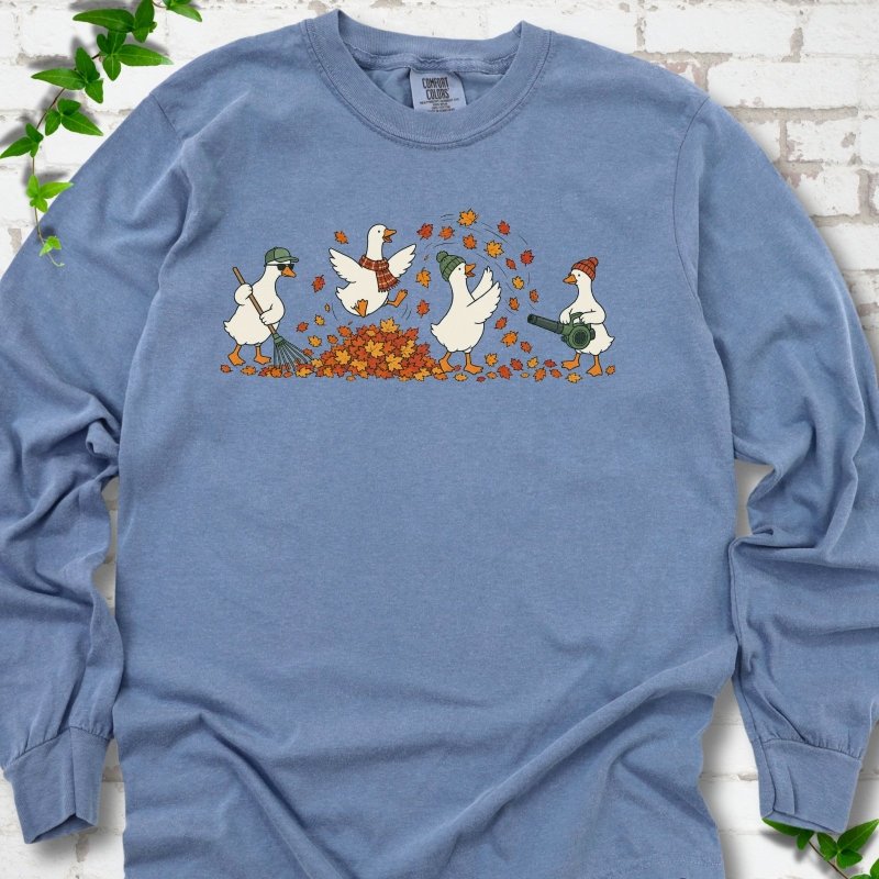 Blue Jean long sleeve T-shirt flat lay with a row of four white cartoon geese doing fall yard work, one raking in a green cap and sunglasses, one leaping into a pile of orange and gold leaves in a plaid scarf, one in a green beanie tossing leaves and one in an orange beanie using a leaf blower, on white brick with ivy sprigs.