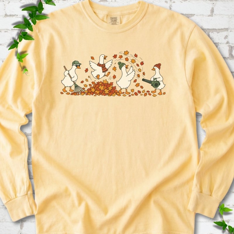 Butter long sleeve T-shirt flat lay with a row of four white cartoon geese doing fall yard work, one raking in a green cap and sunglasses, one leaping into a pile of orange and gold leaves in a plaid scarf, one in a green beanie tossing leaves and one in an orange beanie using a leaf blower, on white brick with ivy sprigs.
