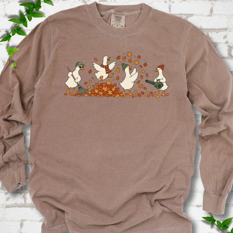 Espresso long sleeve T-shirt flat lay with a row of four white cartoon geese doing fall yard work, one raking in a green cap and sunglasses, one leaping into a pile of orange and gold leaves in a plaid scarf, one in a green beanie tossing leaves and one in an orange beanie using a leaf blower, on white brick with ivy sprigs.