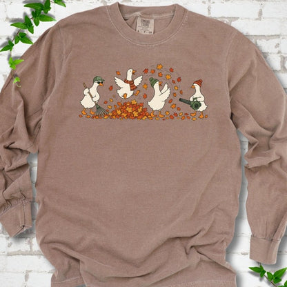 Espresso long sleeve T-shirt flat lay with a row of four white cartoon geese doing fall yard work, one raking in a green cap and sunglasses, one leaping into a pile of orange and gold leaves in a plaid scarf, one in a green beanie tossing leaves and one in an orange beanie using a leaf blower, on white brick with ivy sprigs.