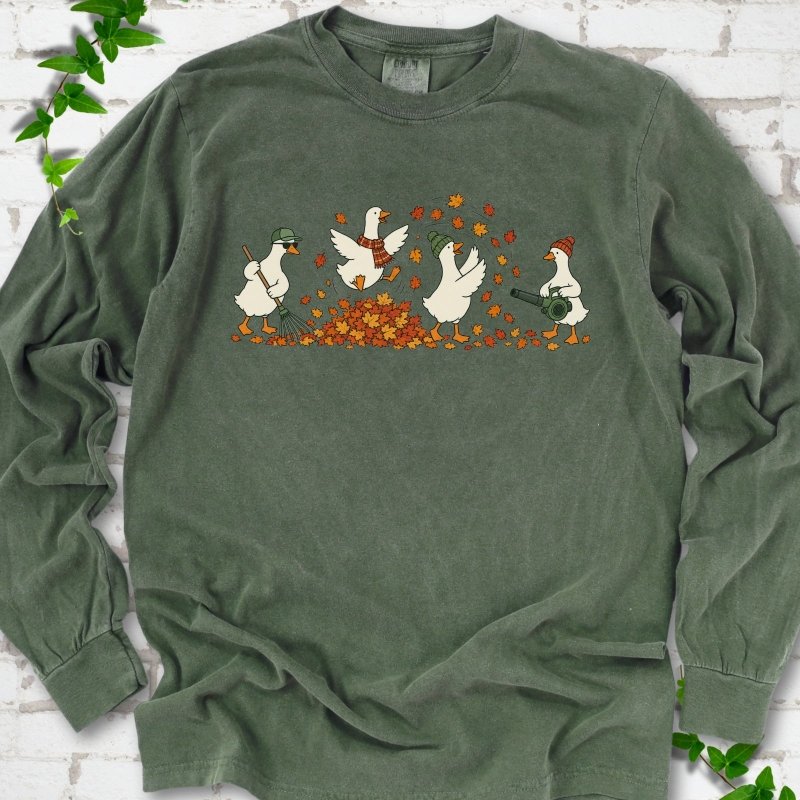 Hemp long sleeve T-shirt flat lay with a row of four white cartoon geese doing fall yard work, one raking in a green cap and sunglasses, one leaping into a pile of orange and gold leaves in a plaid scarf, one in a green beanie tossing leaves and one in an orange beanie using a leaf blower, on white brick with ivy sprigs.