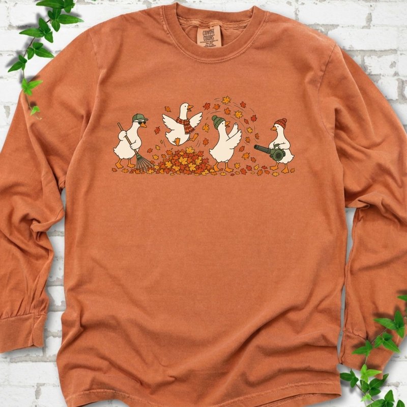 Yam long sleeve T-shirt flat lay with a row of four white cartoon geese doing fall yard work, one raking in a green cap and sunglasses, one leaping into a pile of orange and gold leaves in a plaid scarf, one in a green beanie tossing leaves and one in an orange beanie using a leaf blower, on white brick with ivy sprigs.