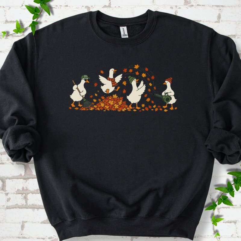Black sweatshirt flat lay with a row of four white cartoon geese doing fall yard work, one raking in a green cap and sunglasses, one leaping into a pile of orange and gold leaves in a plaid scarf, one in a green beanie tossing leaves and one in an orange beanie using a leaf blower, on white brick with ivy sprigs.