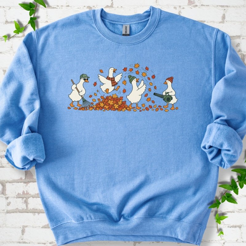 Carolina Blue sweatshirt flat lay with a row of four white cartoon geese doing fall yard work, one raking in a green cap and sunglasses, one leaping into a pile of orange and gold leaves in a plaid scarf, one in a green beanie tossing leaves and one in an orange beanie using a leaf blower, on white brick with ivy sprigs.