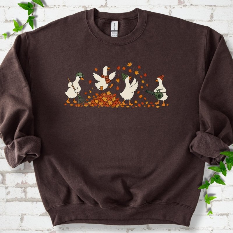 Dark Chocolate sweatshirt flat lay with a row of four white cartoon geese doing fall yard work, one raking in a green cap and sunglasses, one leaping into a pile of orange and gold leaves in a plaid scarf, one in a green beanie tossing leaves and one in an orange beanie using a leaf blower, on white brick with ivy sprigs.