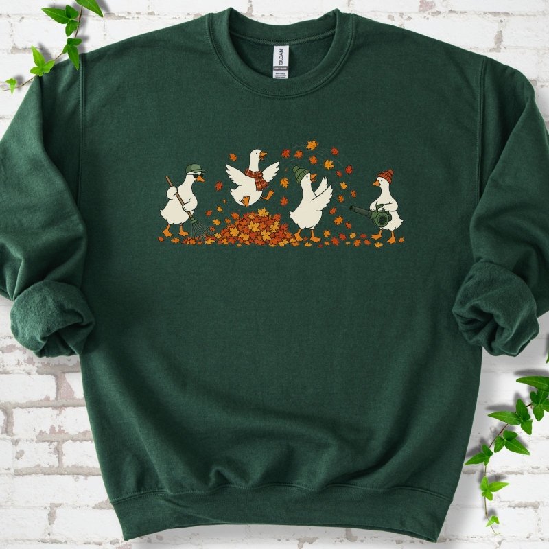 Forest Green sweatshirt flat lay with a row of four white cartoon geese doing fall yard work, one raking in a green cap and sunglasses, one leaping into a pile of orange and gold leaves in a plaid scarf, one in a green beanie tossing leaves and one in an orange beanie using a leaf blower, on white brick with ivy sprigs.