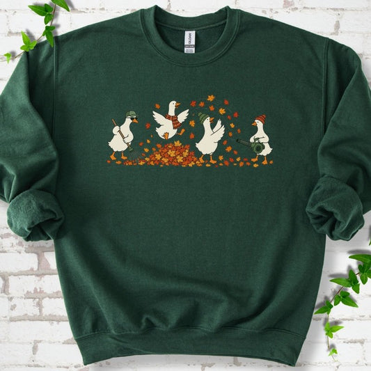 Forest Green sweatshirt flat lay with a row of four white cartoon geese doing fall yard work, one raking in a green cap and sunglasses, one leaping into a pile of orange and gold leaves in a plaid scarf, one in a green beanie tossing leaves and one in an orange beanie using a leaf blower, on white brick with ivy sprigs.