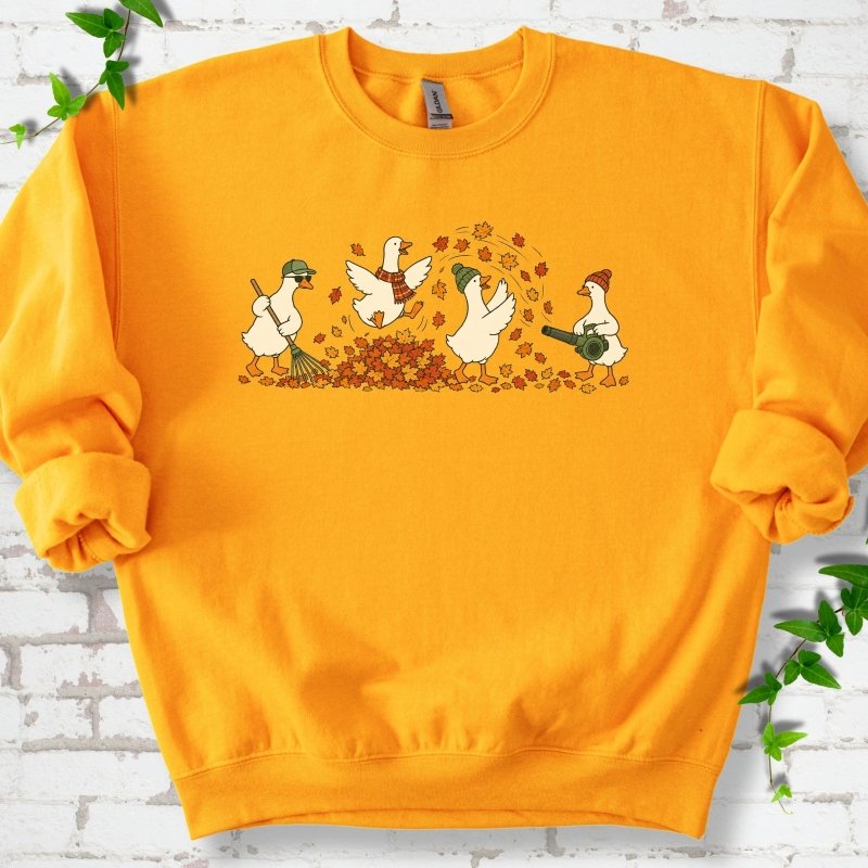 Gold sweatshirt flat lay with a row of four white cartoon geese doing fall yard work, one raking in a green cap and sunglasses, one leaping into a pile of orange and gold leaves in a plaid scarf, one in a green beanie tossing leaves and one in an orange beanie using a leaf blower, on white brick with ivy sprigs.